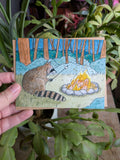 A greeting card with watercolour art of a raccoon sitting by a campfire toasting a marshmallow.